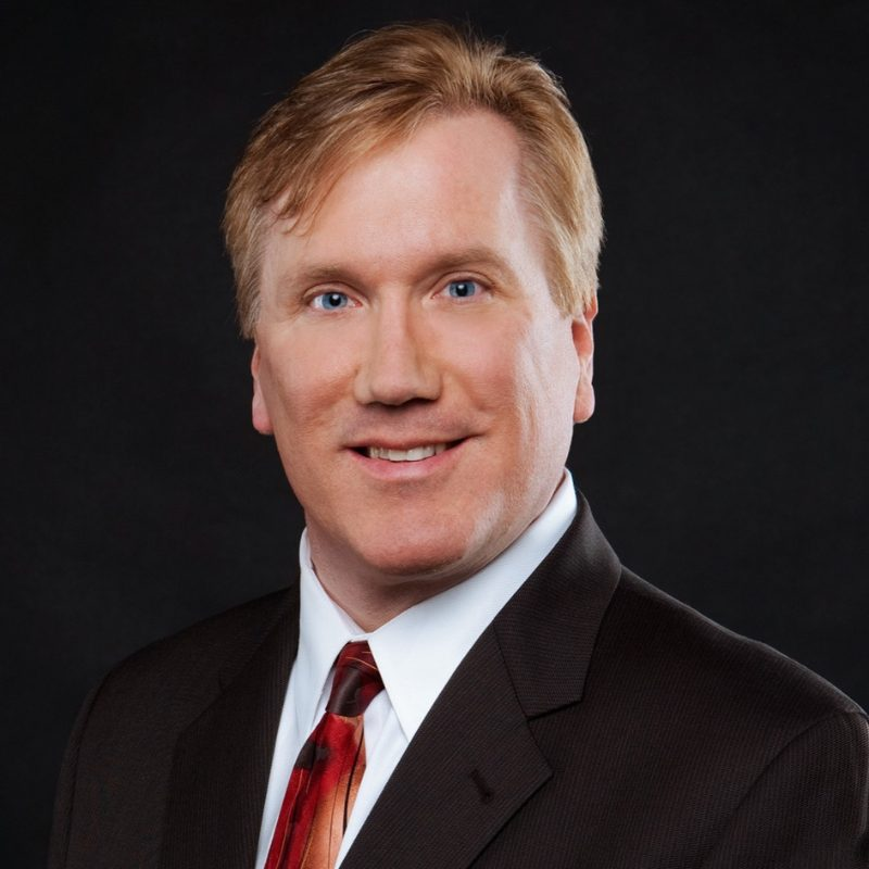 Professional head-and-shoulders portrait of a smiling man in a dark suit and red tie against a black background.
